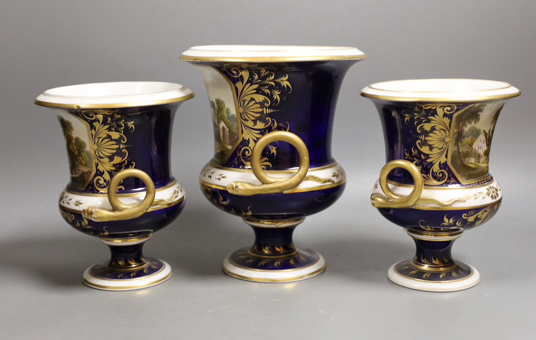 A garniture of three Derby named twin handled cups, View near Belper, Derbyshire, and 'View in North Wales' and 'View near Rome', 17cm and 20.5cm
